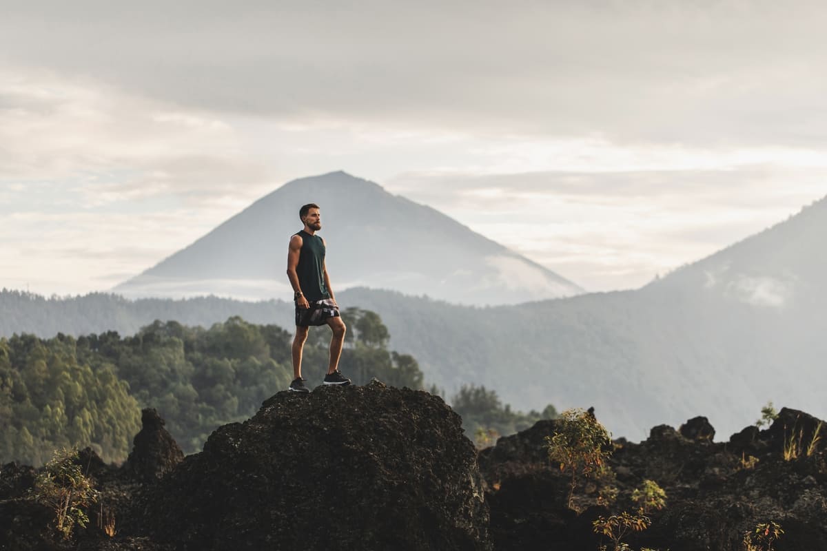 Pemuda itu berdiri di atas batu dan menikmati pemandangan gunung berapi Agung yang menakjubkan di Bali. Konsep perjalanan dan gaya hidup aktif.