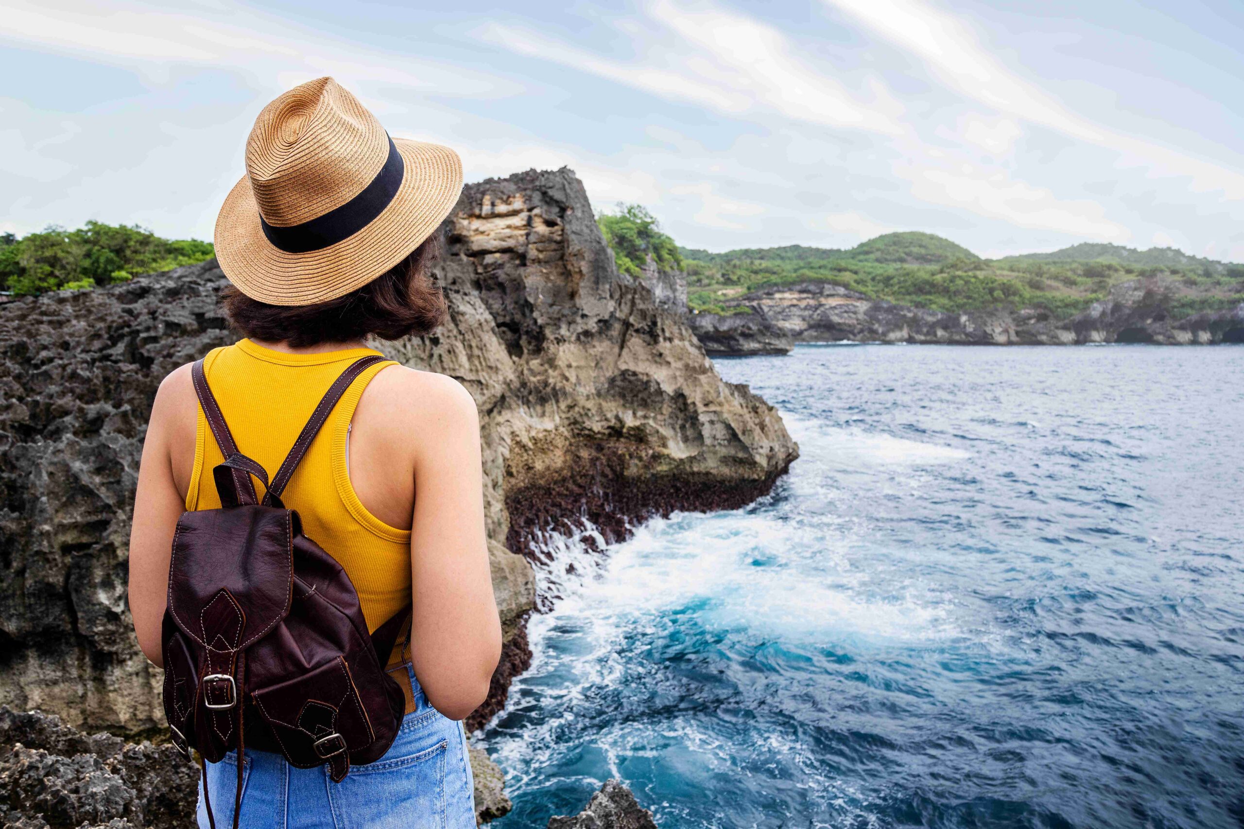 Rear view of woman with hat and backpack contemplating nature. Female looking at ocean view. Angel’s Billabong, Bali.