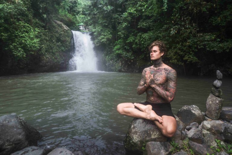 tattooed man sitting in lotus position on rock with Aling Aling waterfall and green plants on