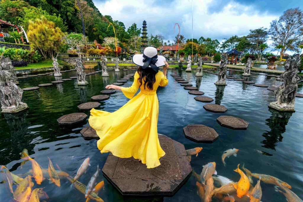 Woman with a yellow dress standing in a pond, colorful fish at Tirta Gangga Water Palace in Bali