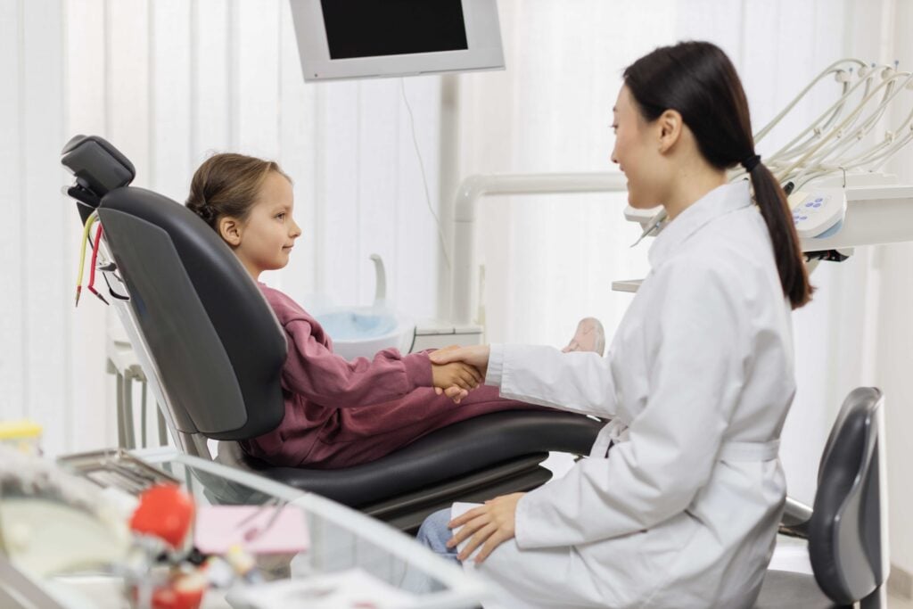 Small caucasian satisfied smiling child patient shakes asian dentist's hand