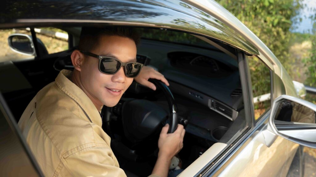 Happy young Asian man in sunglasses driving car in countryside road while traveling alone