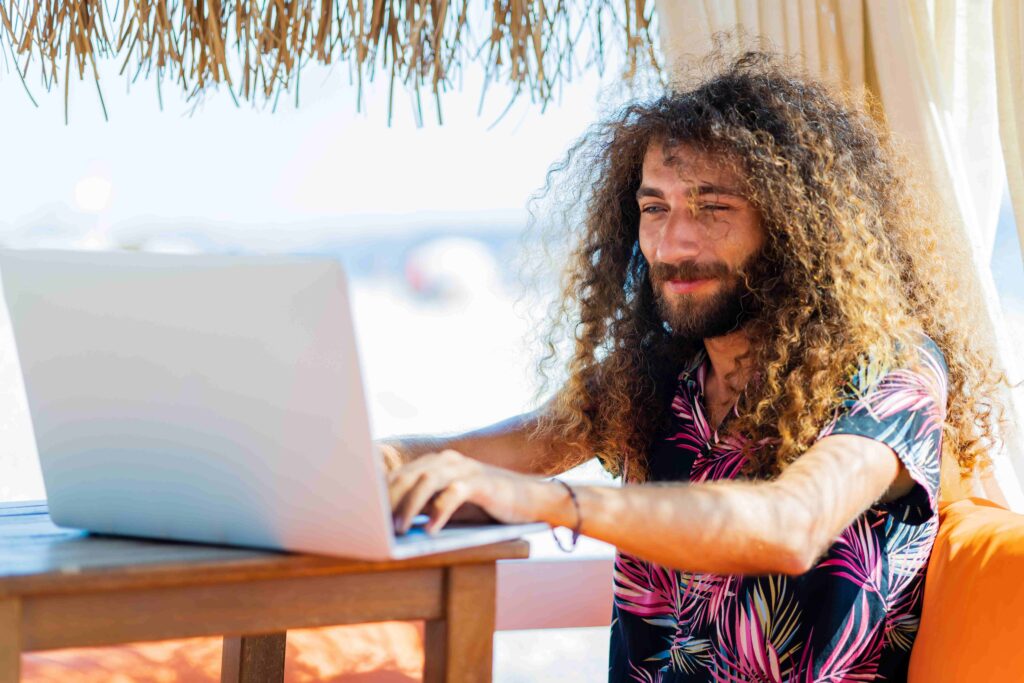 long curly haired man sitting in beach and working laptop vacation