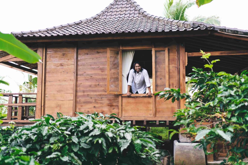Smiling ethnic man standing at window of guest house in tropical garden
