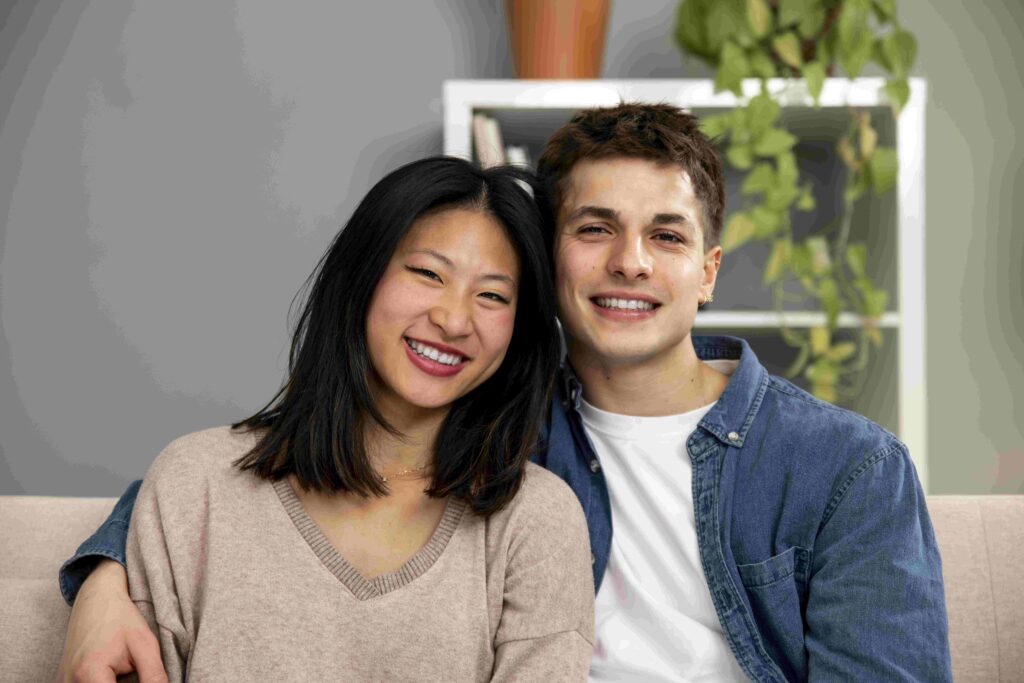 Affectionate smiling young adult couple sitting on sofa at home looking at camera Carefree man and woman relaxing together in couch at the living room