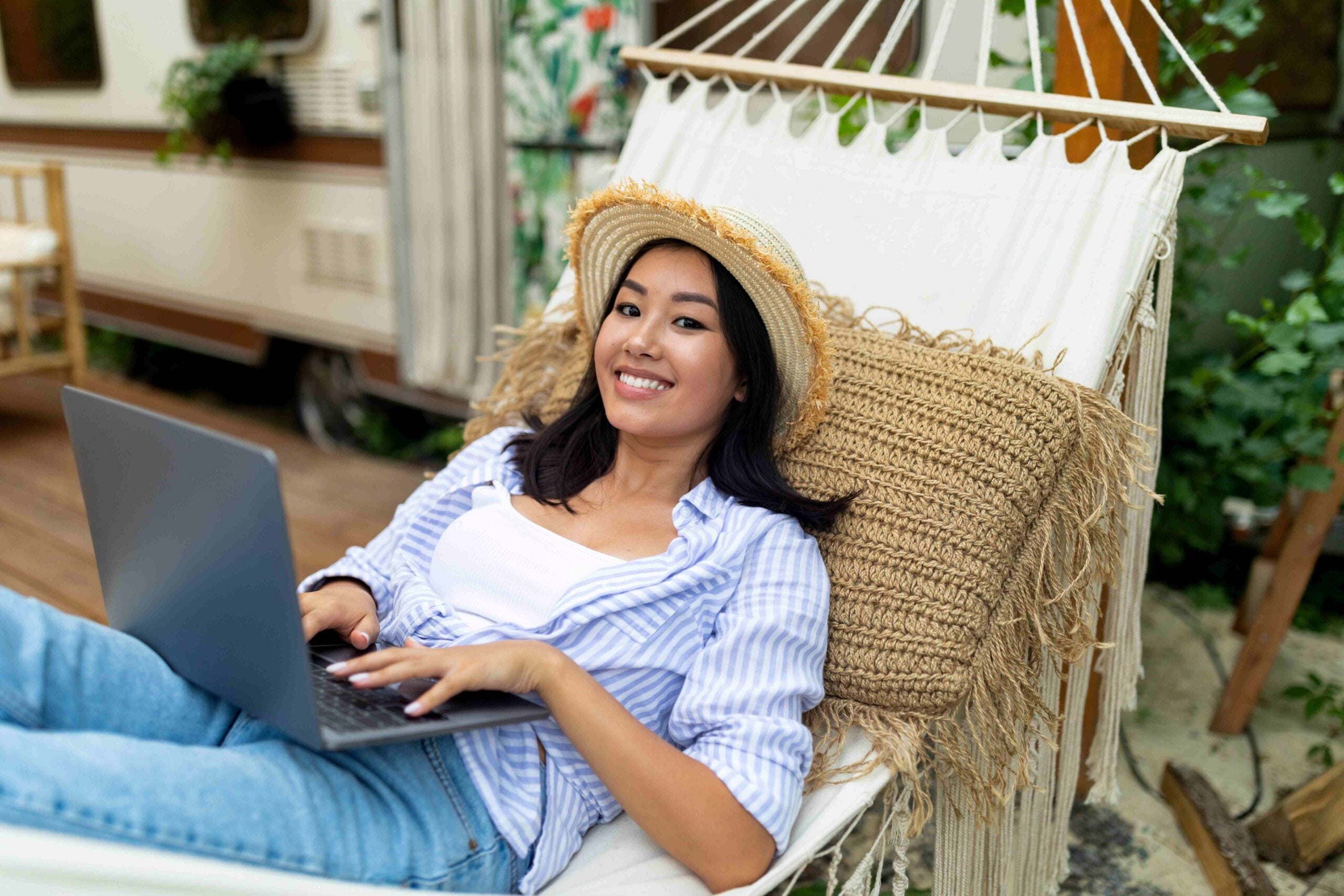 Joyful Asian lady using laptop computer while lying in hammock near RV on autumn camping trip