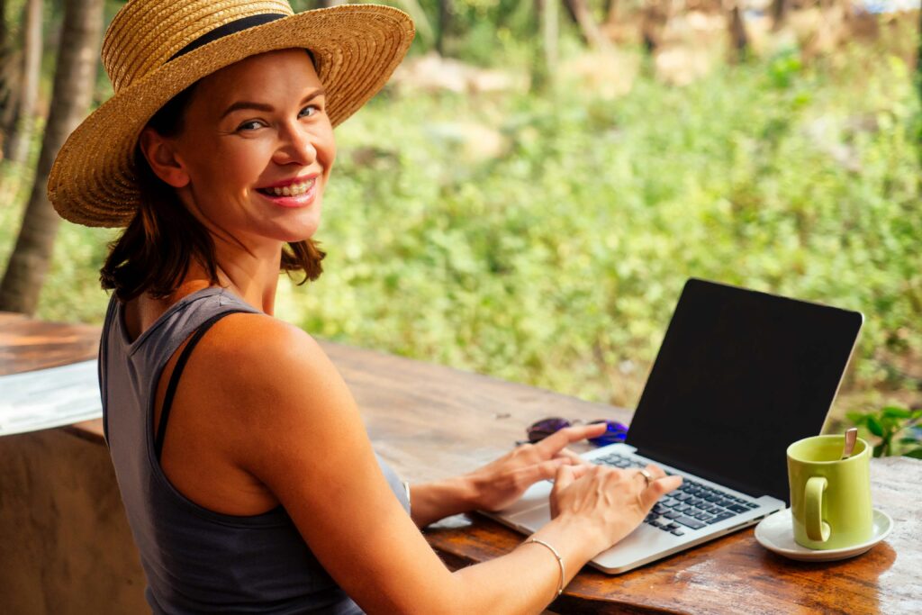 Technology and travel Working outdoors Freelance concept Pretty young woman in hat using laptop in cafe on tropical beach Hooray victory success and successful deal concept promotion at work