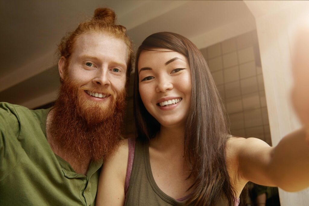 Young good looking hipster man wearing casual shirt posing together with his best female friend with long dark hair while taking self portrait using gadget or electronic device sitting at restaurant