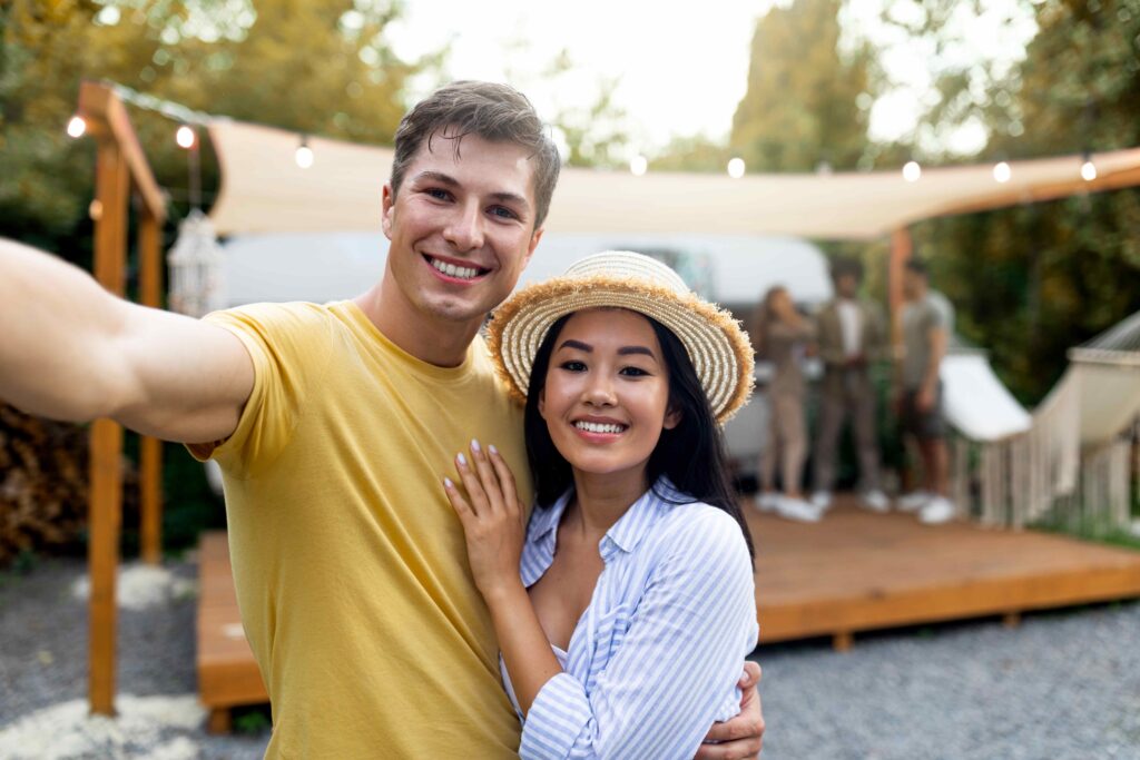 Young happy diverse couple taking selfie at camp, resting with friends near trailer, spending autumn weekend outdoors