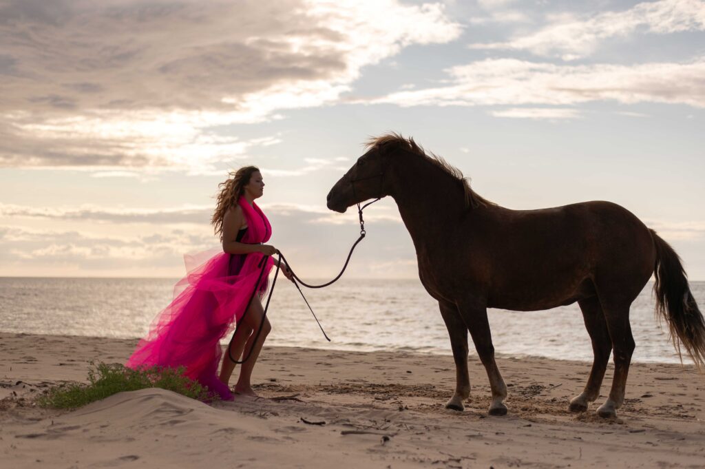 a woman in pink dress standing next to horse on beach