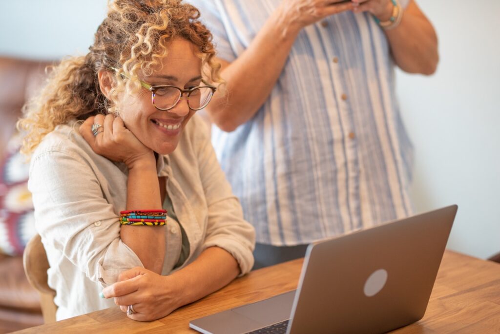 Beautiful blonde curly woman with eyeglasses browsing on laptop at home desk smiling while her mother look at her