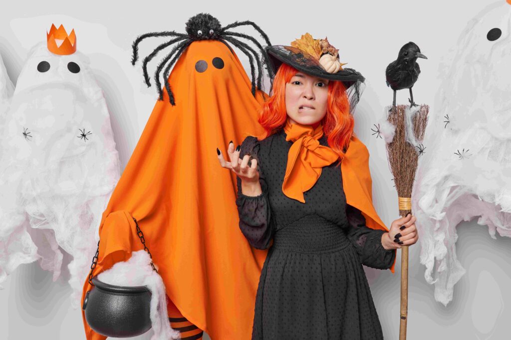 Displeased young woman with orange hair witch wears hat and black dress bites lips feels annoyed holds broom with crow poses together with orange ghost celebrate Halloween pose indoor Creepy carnival