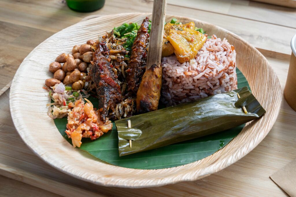 Top down view of a plate of Balinese food with fish, sate, soup, rice, sambal, greens on bamboo plate