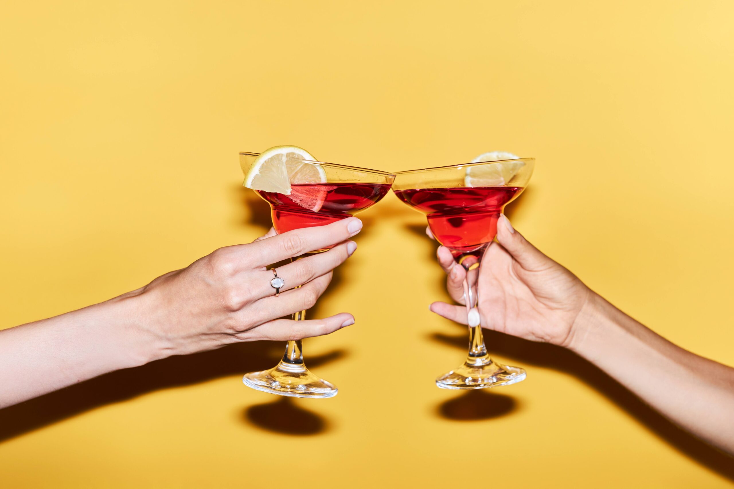 Close up of two female hands holding red cocktails against yellow background