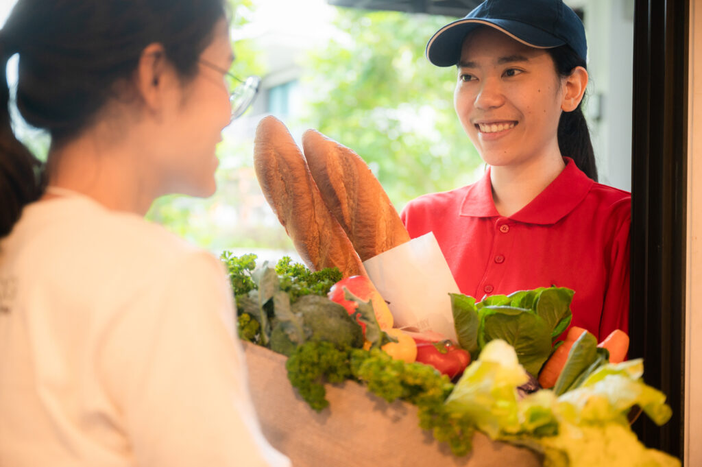 Asian deliver woman in red uniform handling bag of food, fruit, vegetable give to costumer in front of the house, delivery service concept